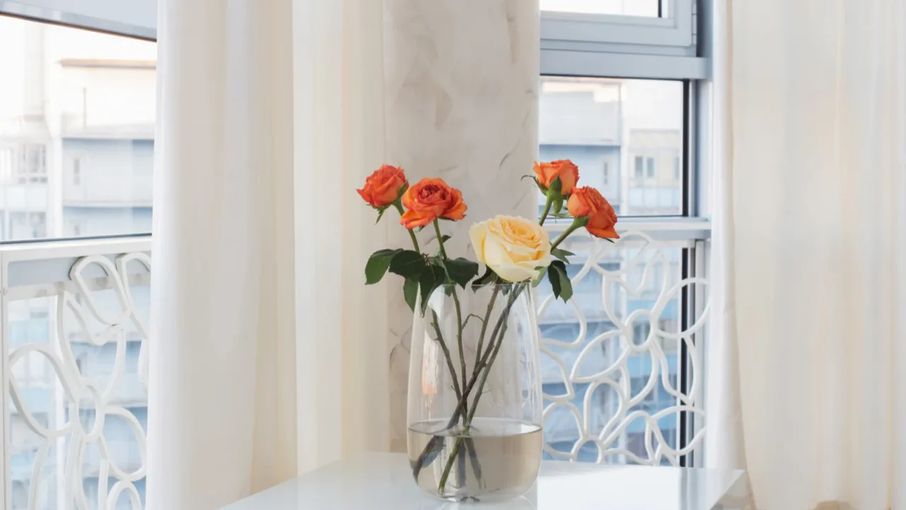 Orange and white roses in vase on a white table in front of a window with sheer white curtains and a window.