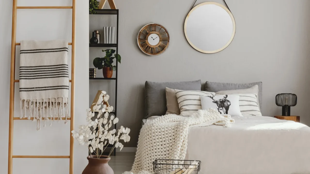 A cozy bedroom interior with knitted throw blanket on bed, shelf with books and decor accents, round decorative mirror, wall clock, flowers in a vase, and wooden ladder with throw blanket.