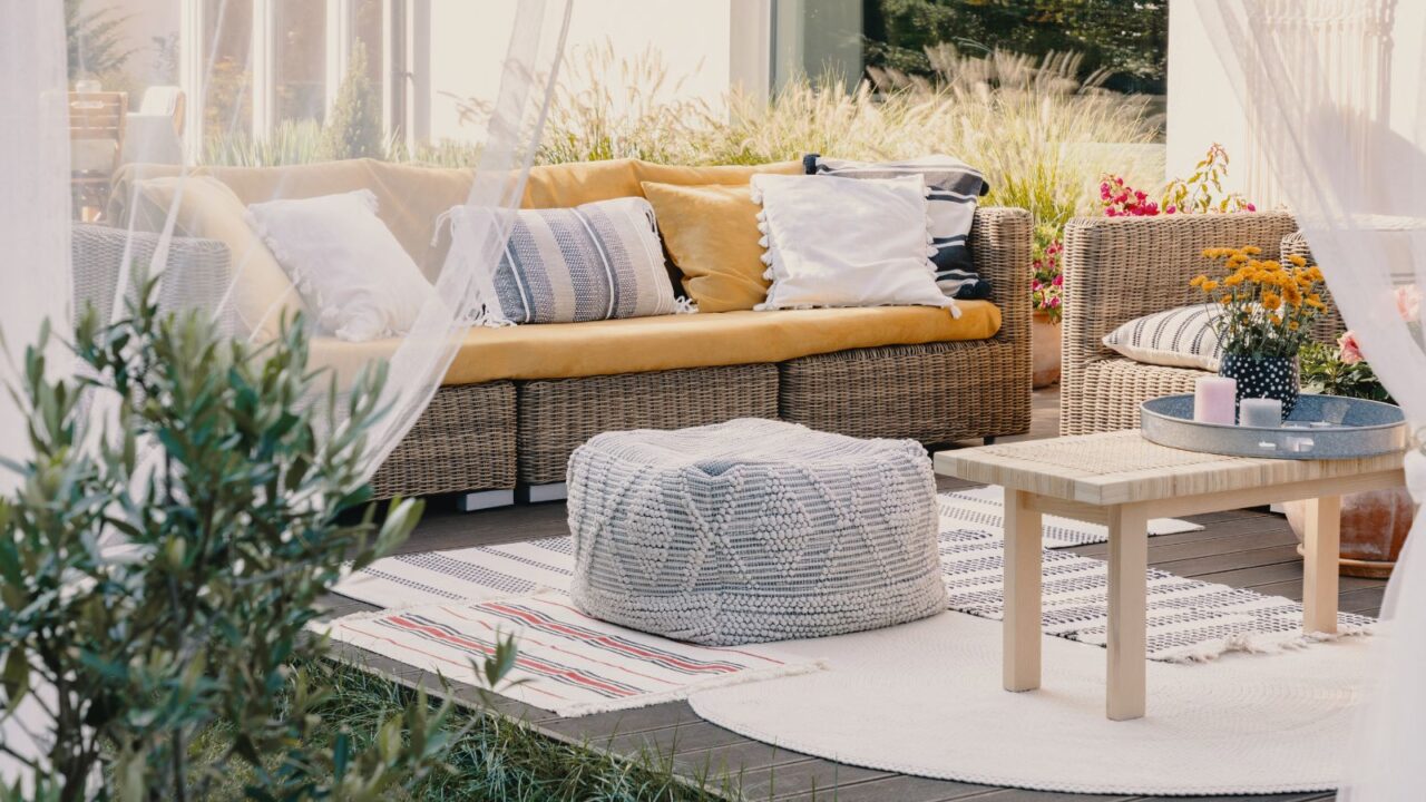 Terrace design with rattan garden furniture set. Cozy yellow, white, black and blue cushions. A centretable with a tray with candles and vase on it. Rugs layered on the floor with a pouf..