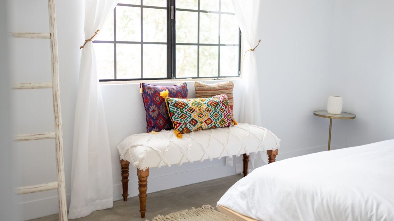 Southwestern Interior design. White bedroom with Southwestern patterned pillows on a white decorative stool in front of a window with white sheer curtains. On the left is a ladder and on the right is a small table.