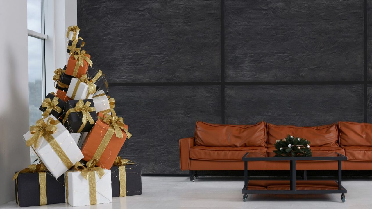 Stylish leather sofa against a black wall, a designer Christmas tree made of boxes with gifts by the window, a table with a Christmas wreath in a spacious room. Minimalism. Stylish Christmas interior.