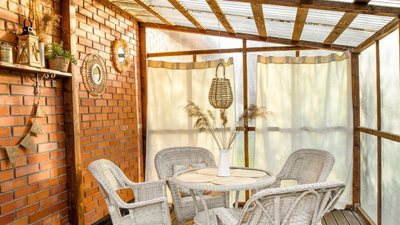 Boho style outdoor decor with curtains and wicker, rattan, and reed decorations. Wall made with greenhouse plastic and ceiling is polycarbonate. White garden furniture and mirrors on brick wall.