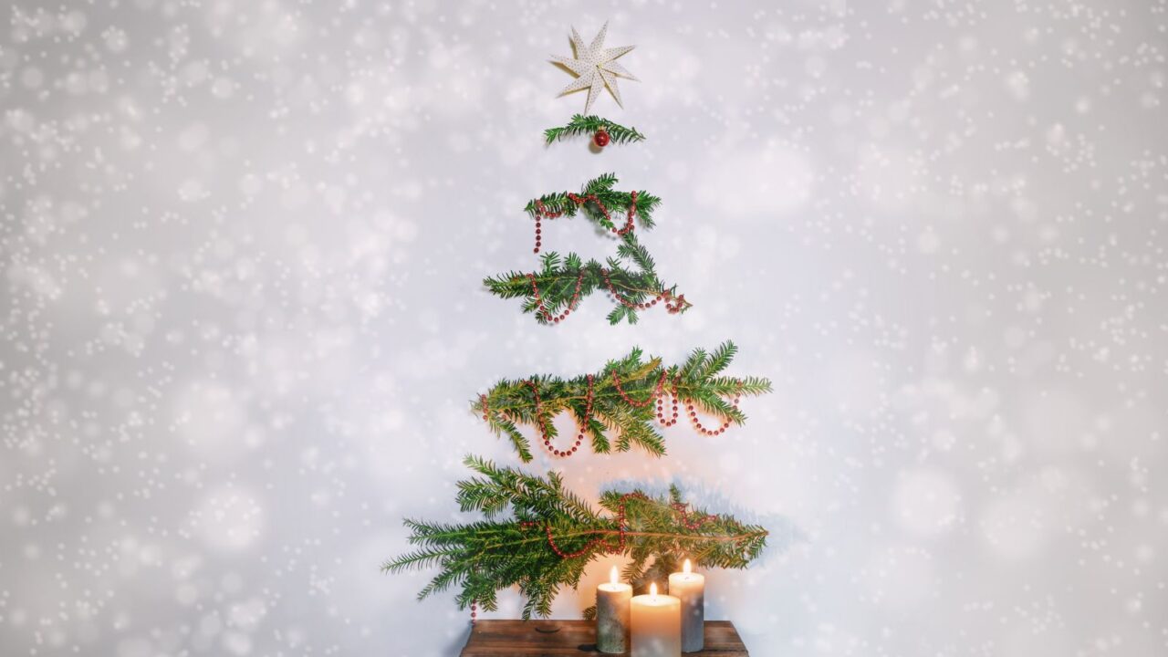 Christmas tree alternative. Evergreen branches with decoration hanging on the wall above a rustic wooden stool with candles. Sustainable holidays against a snowy background.