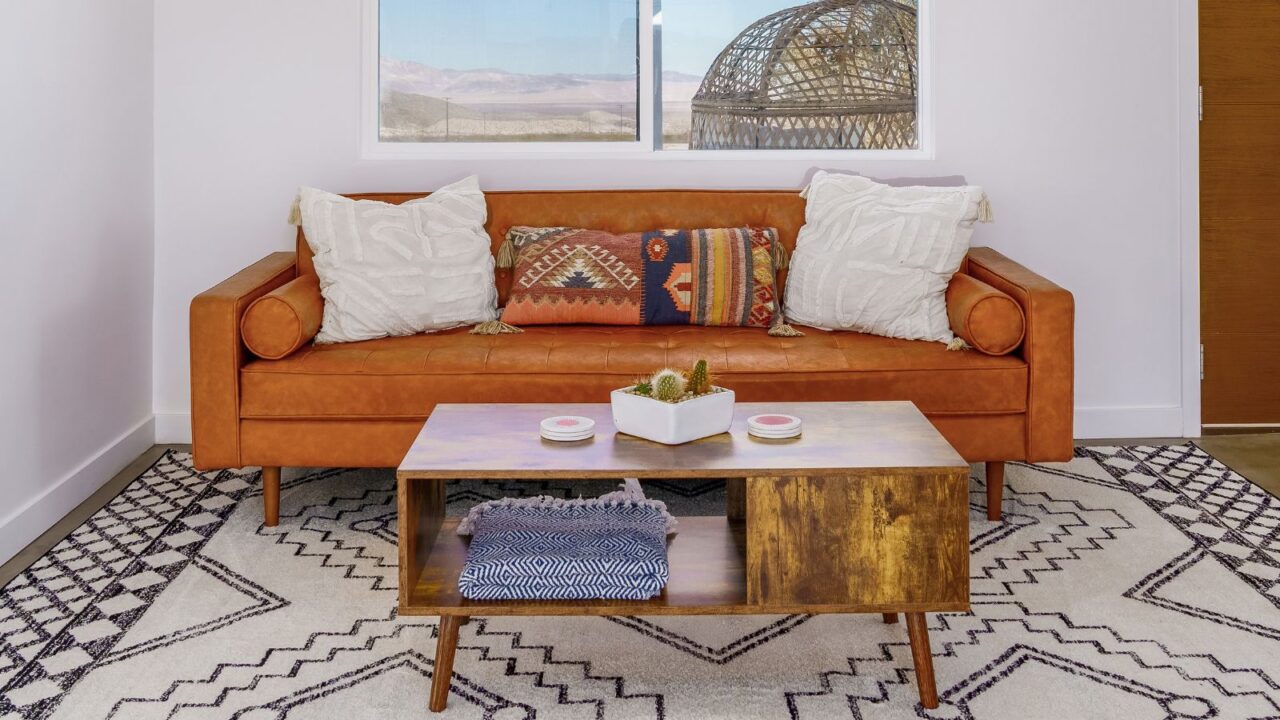 A living room with leather couch with cushions with Southwest patterns and a designer Southwest rug. A rustic coffee table in the centre table with a cactus on the table and a window at the back.