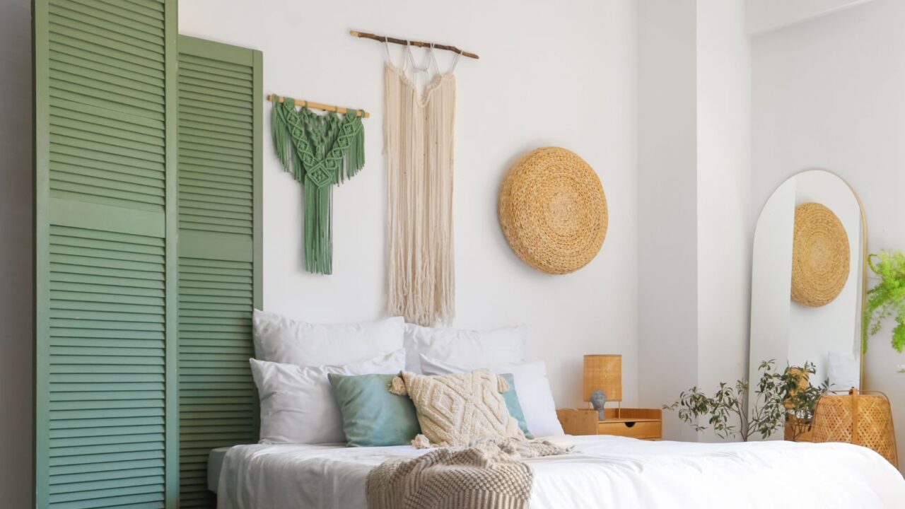 A bedroom interior with a cozy bed with throw blanket and pillows, floor standing mirror, and woven wall hangings.