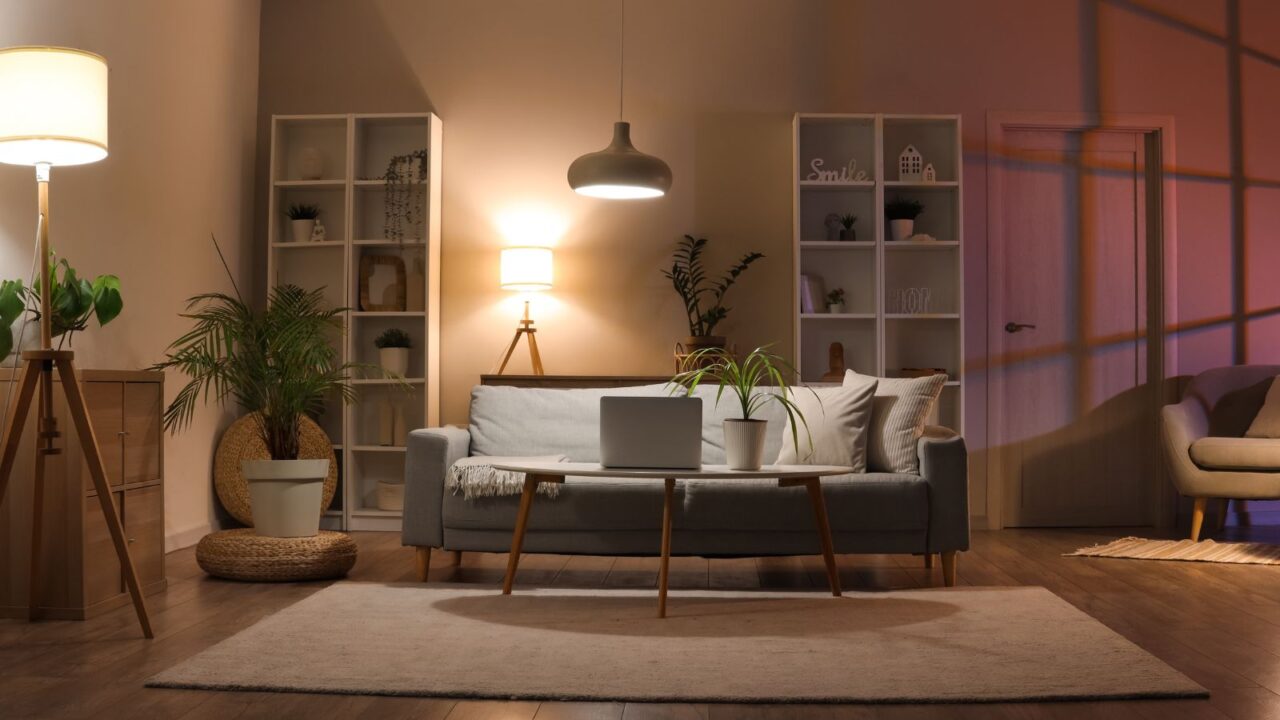 Interior of living room with sofa, armchair, shelves, a rattan pouf with a plant on top, table, hanging light, table lamp, and floor lamp. Soft glowing lamps in evening/night time.