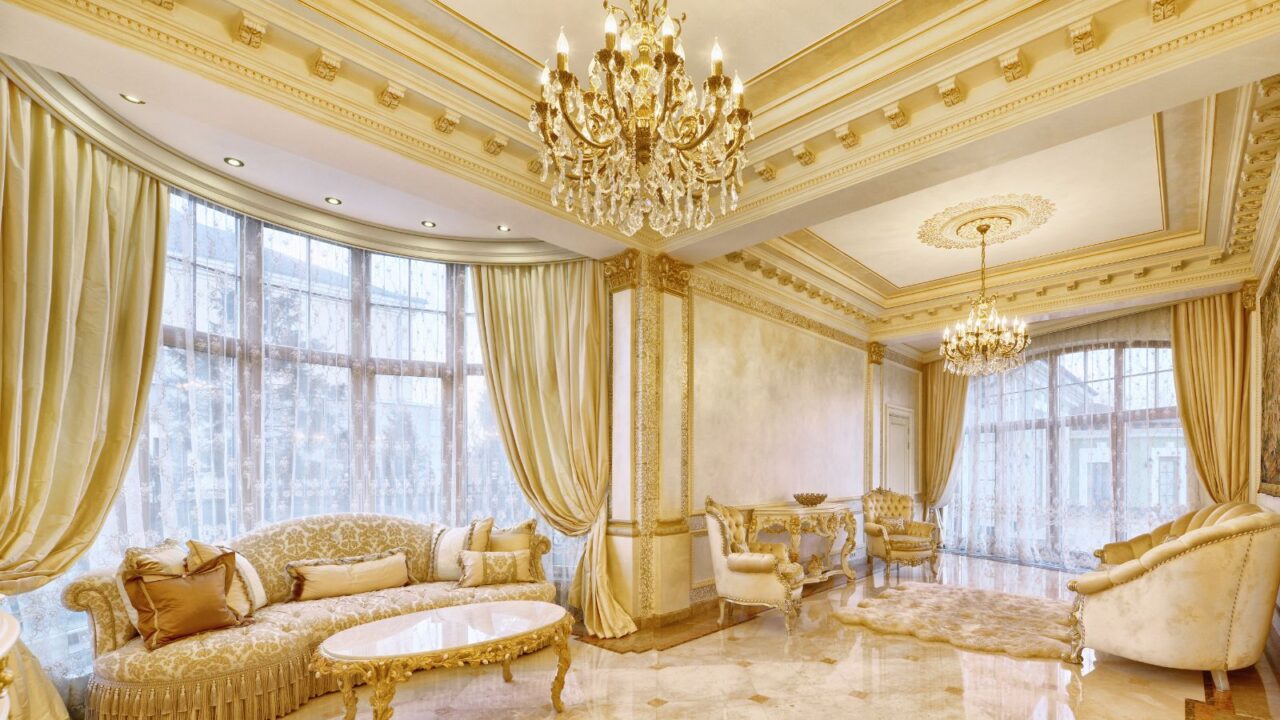 A royal looking while and gold drawing room. Large windows with curtains, sofa sets, furniture, and a chandelier. Golden metallic finishes on the ceiling.