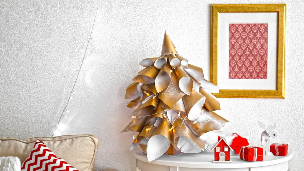 DIY handmade paper Christmas tree on a white table with ribbons, lights, and stars. Red and white Christmas decor on the table. String lights are hanging on the wall. A gold frame hanging on the wall. A red and white pattern cushion on an off white sofa.