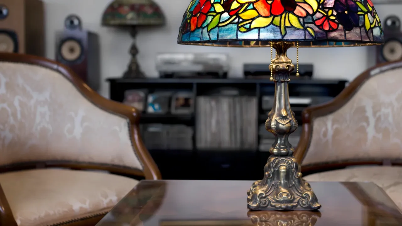 Antique stained glass lamp on a table. In the background, there are two upholstered chairs and a shelf displaying various items.