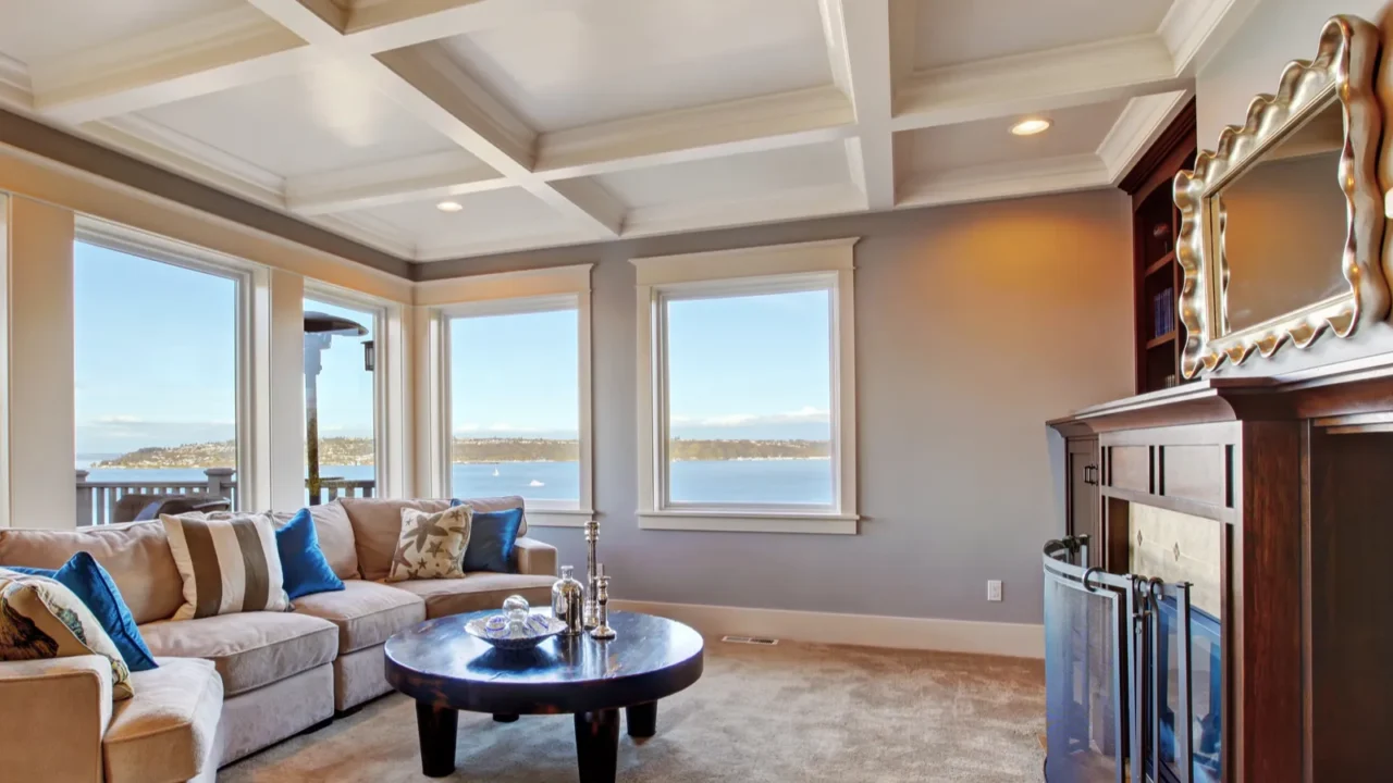 Warm living room interior in luxury house with coffered white ceiling, big windows facing sea, a sofa set and a table.