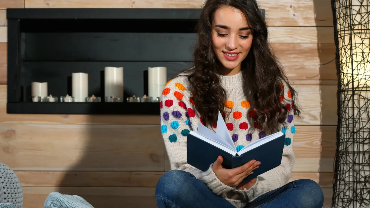 young beautiful woman in warm sweater reading book on rug