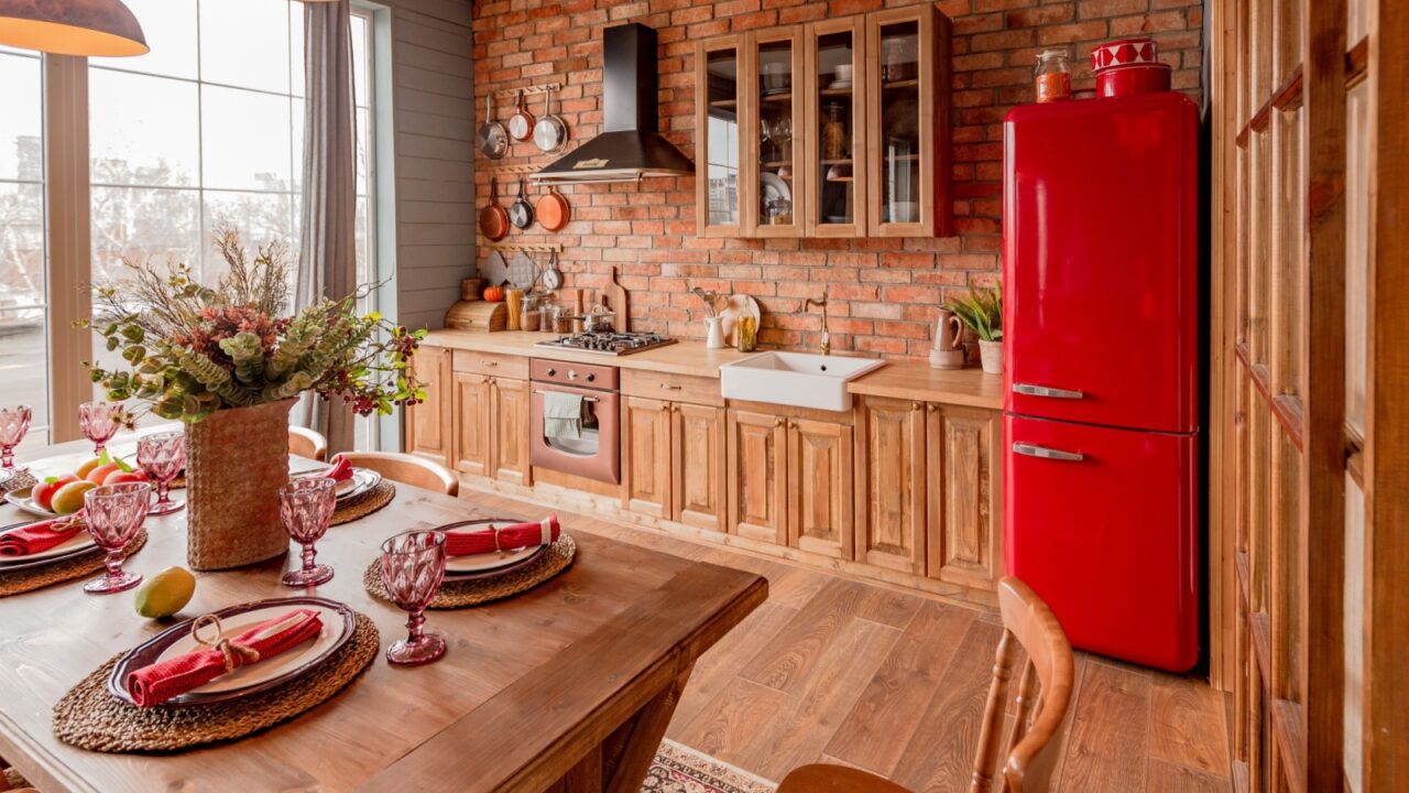 A rustic kitchen with a red refrigerator, wooden cabinets, and a brick backsplash.