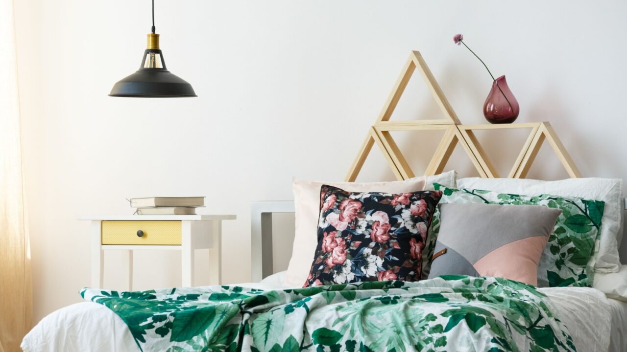 A bedroom interior with bed with floral pattern pillows and blanket, statement ceiling light, and sleek white nightstand with books on it.