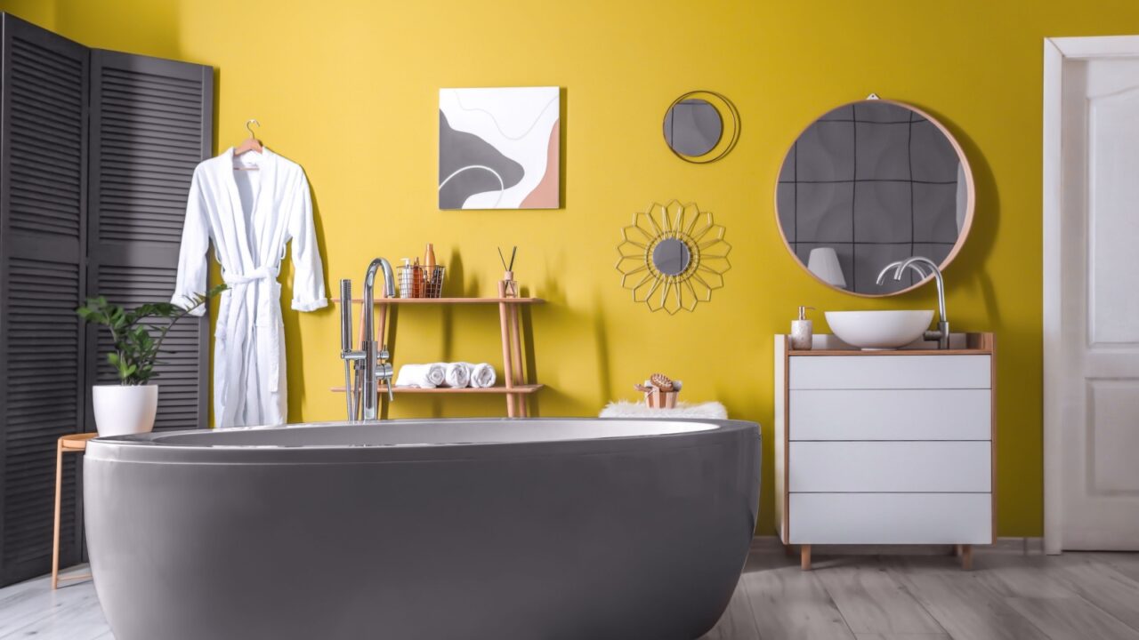 A modern bathroom with a yellow wall, a gray bathtub, a potted plant, a wooden table, shelves, and wall art.