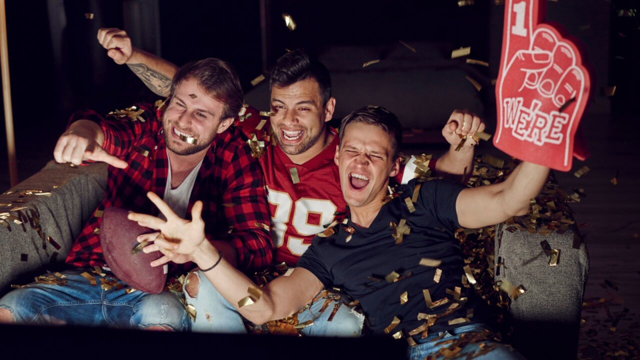 Three male friends celebrating on a couch with confetti, one holding a foam finger and another holding a football.