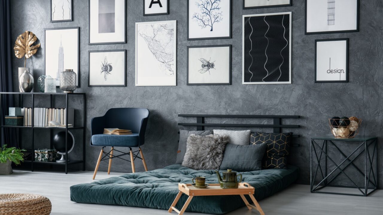 A modern bedroom interior with blue armchair, green futon, framed artwork on wall, and bookcase with books and decor accents.