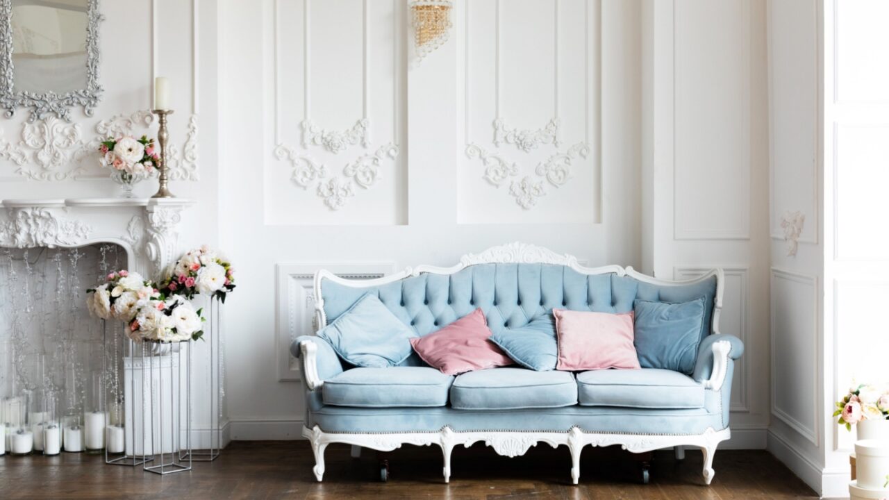 A vintage-style living room with a light blue tufted sofa, pink and blue pillows, ornate white wall moldings, a white fireplace with floral arrangements, and candles.