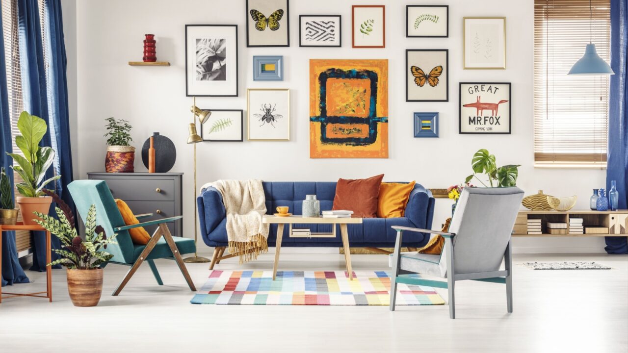 A modern living room with a blue sofa, colorful cushions, a variety of plants, a multi-colored rug, and a gallery wall featuring various framed artworks and prints.