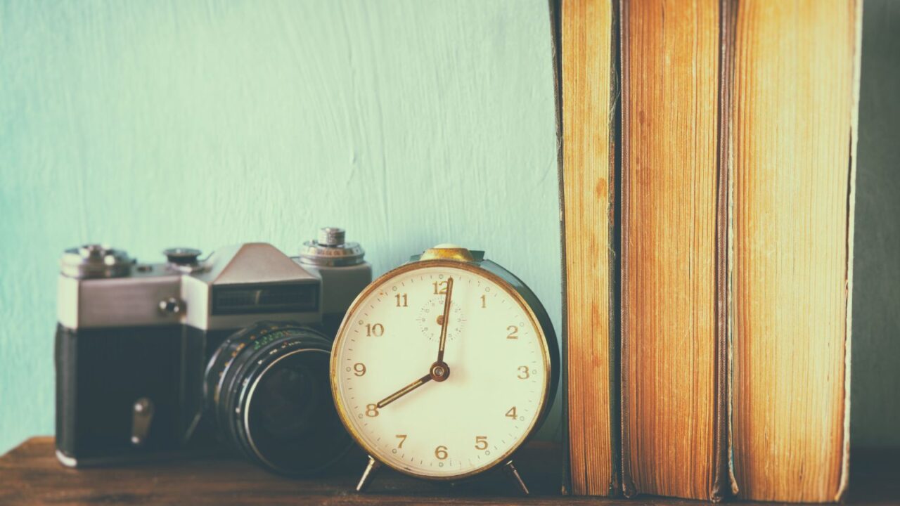 A vintage clock, camera, and books on a wooden shelf.