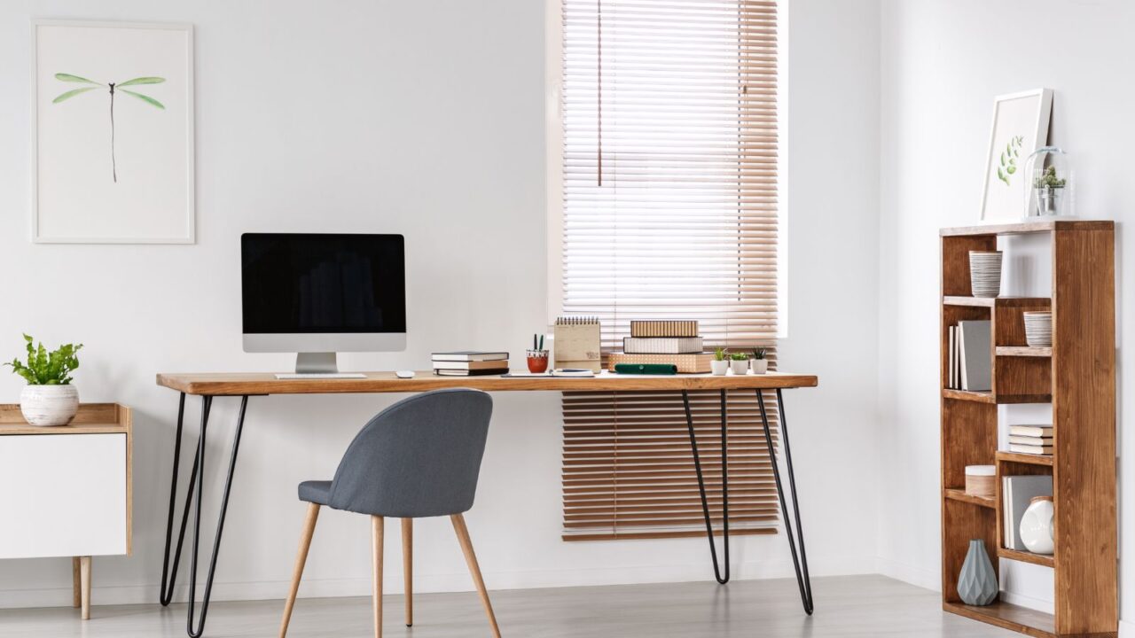 A home office interior with white walls, poster, wooden table, chair, computer, small potted plants, and a wooden bookshelf.