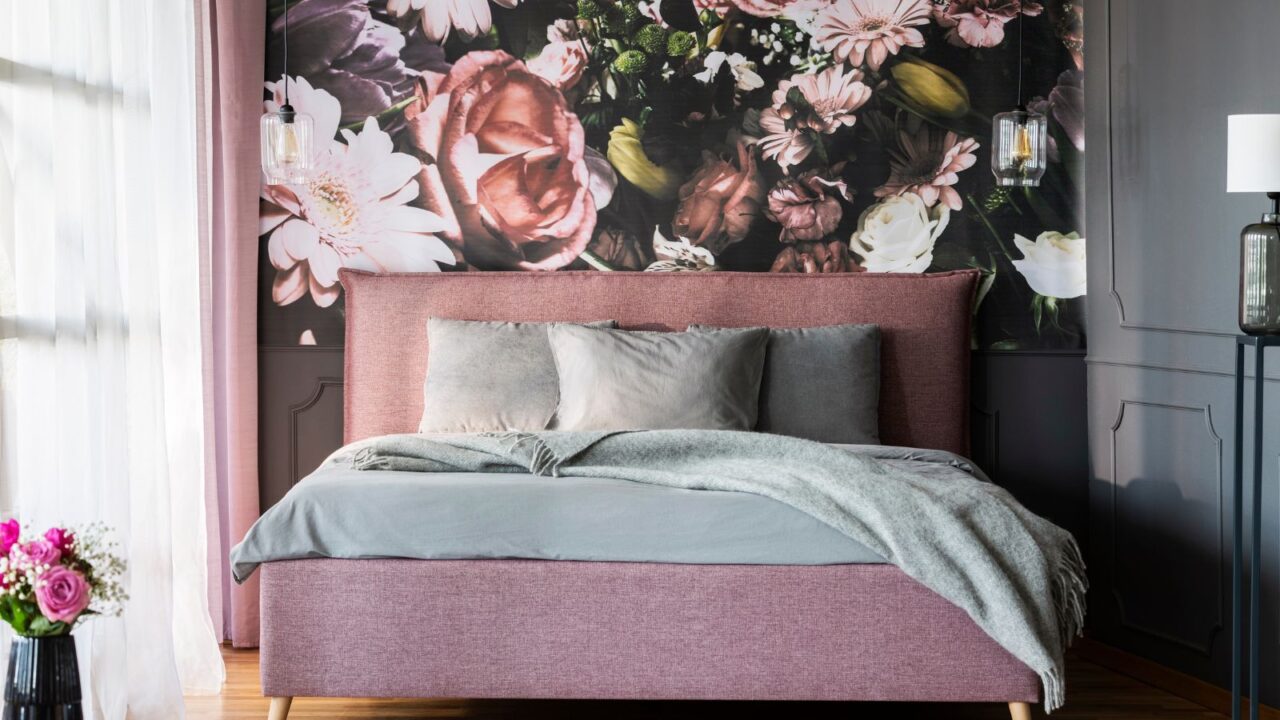 A bedroom interior with a pink bed with throw blanket and pillows, and a floral wallpaper.