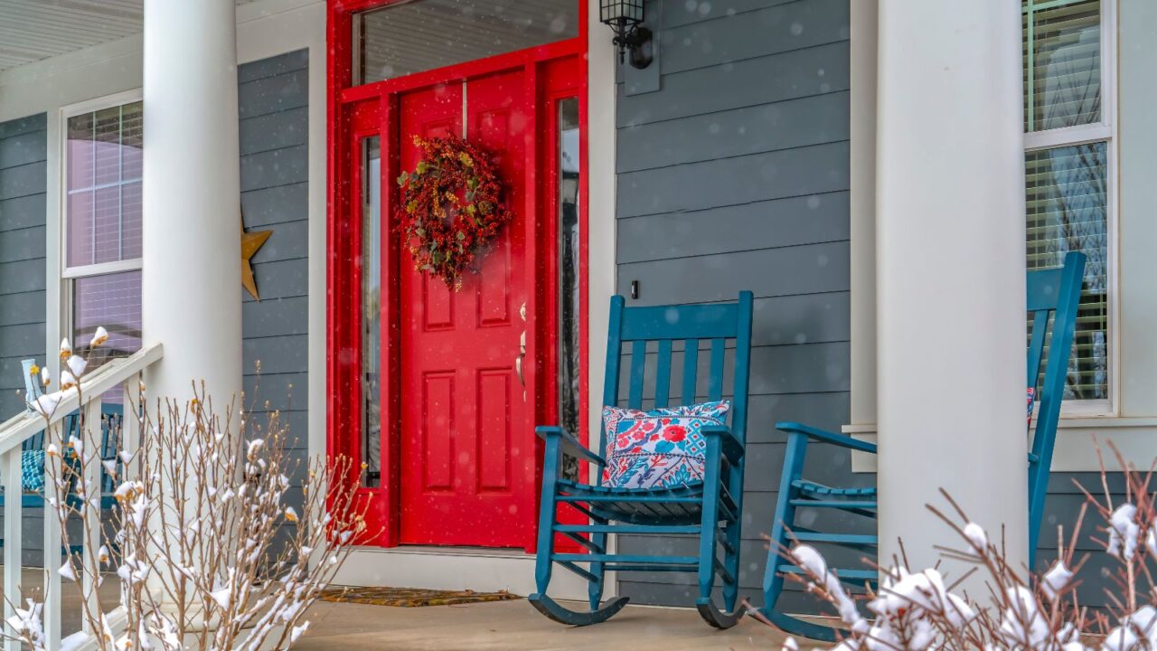 Two blue rocking chairs with cushions on a porch and a fire engine red color door with a wreath on it.