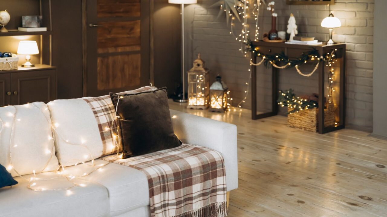 A living room interior with a cozy white sofa with throw blanket, cushion, and string lights, wooden console table, and lanterns.