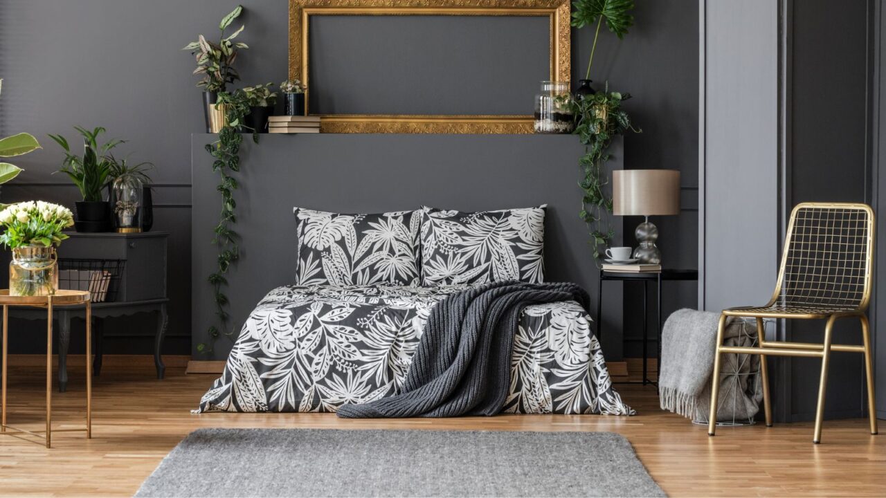 A stylish bedroom interior with metal accent chair, table, plants, and a cozy bed with floral bedding and a throw blanket.