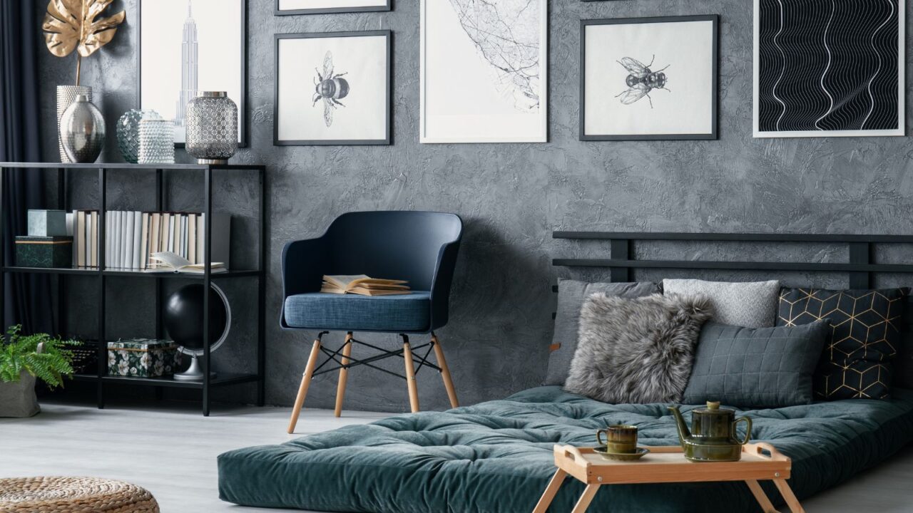 A modern bedroom interior with blue accent chair, green futon, framed artwork on wall, and bookcase with books and decor accents.