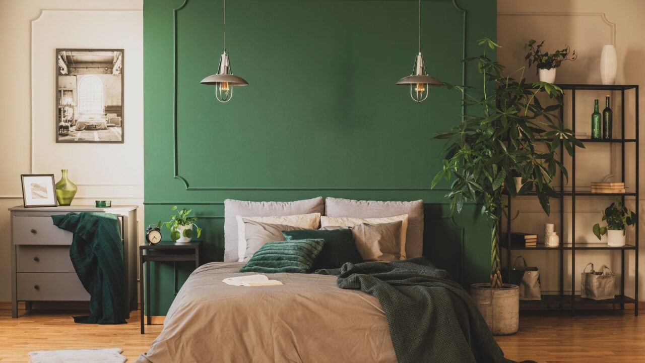 A stylish bedroom interior with a green accent wall, cozy bed, throw blanket, pillows, potted plants, and metallic shelf with decor accents.