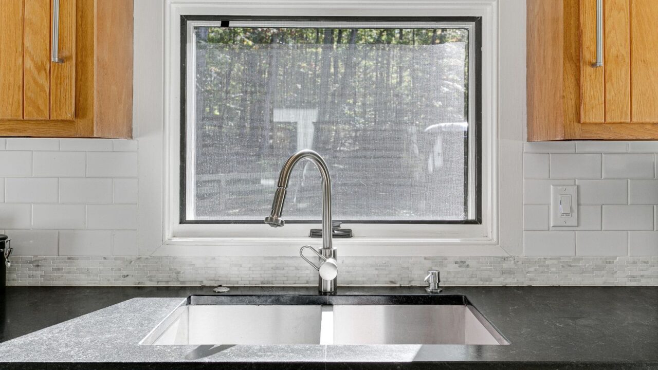 A kitchen interior with wooden cabinets, subway tiles backsplash, marble countertop, and a high arc stainless steel faucet.