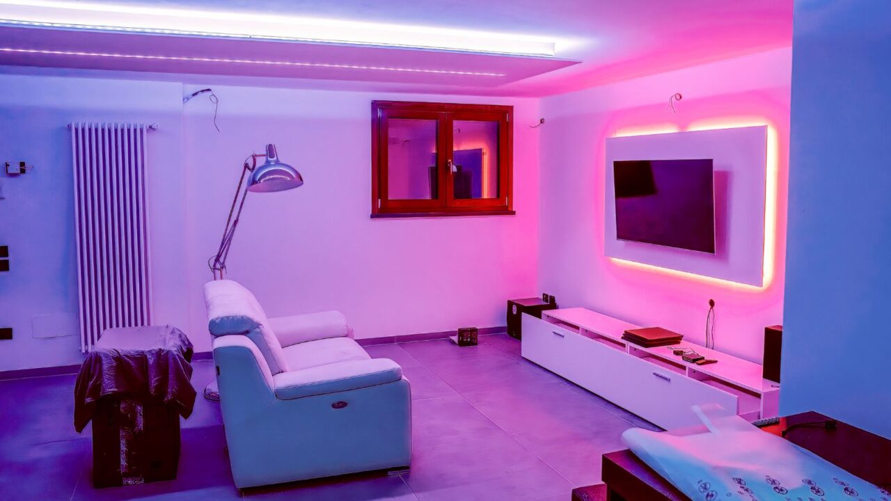 An entertainment room interior with white sofa, floor lamp, smart tv, and led lights in the room.