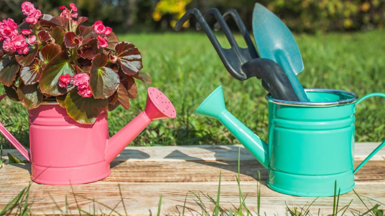 A DIY pink and green watering cans filled with flowers and gardening tools in them.