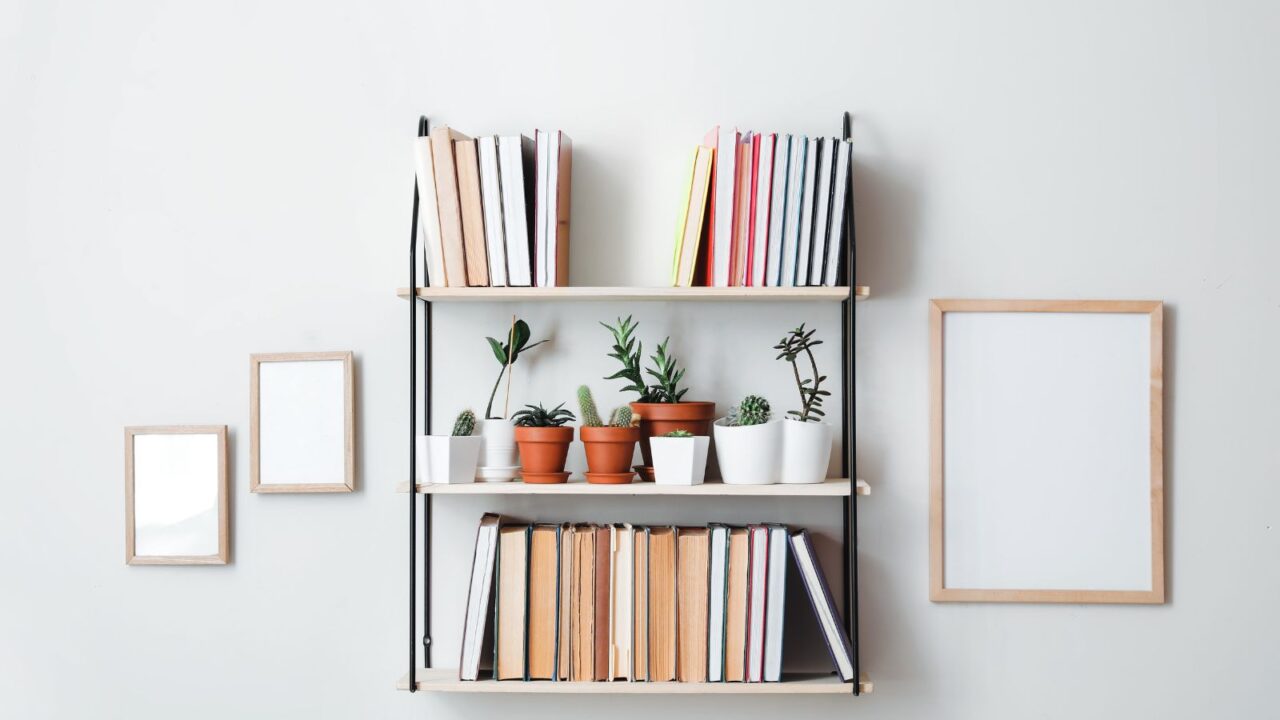 A wall mounted bookshelf with books and small potted plants and succulents, and empty wooden frames on the wall.