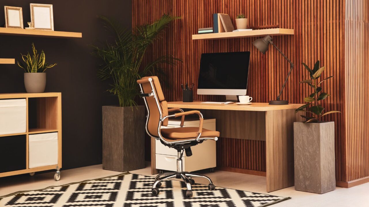 A home office interior with a wooden table, chair, desktop, lamp, floating shelf, and a rug.