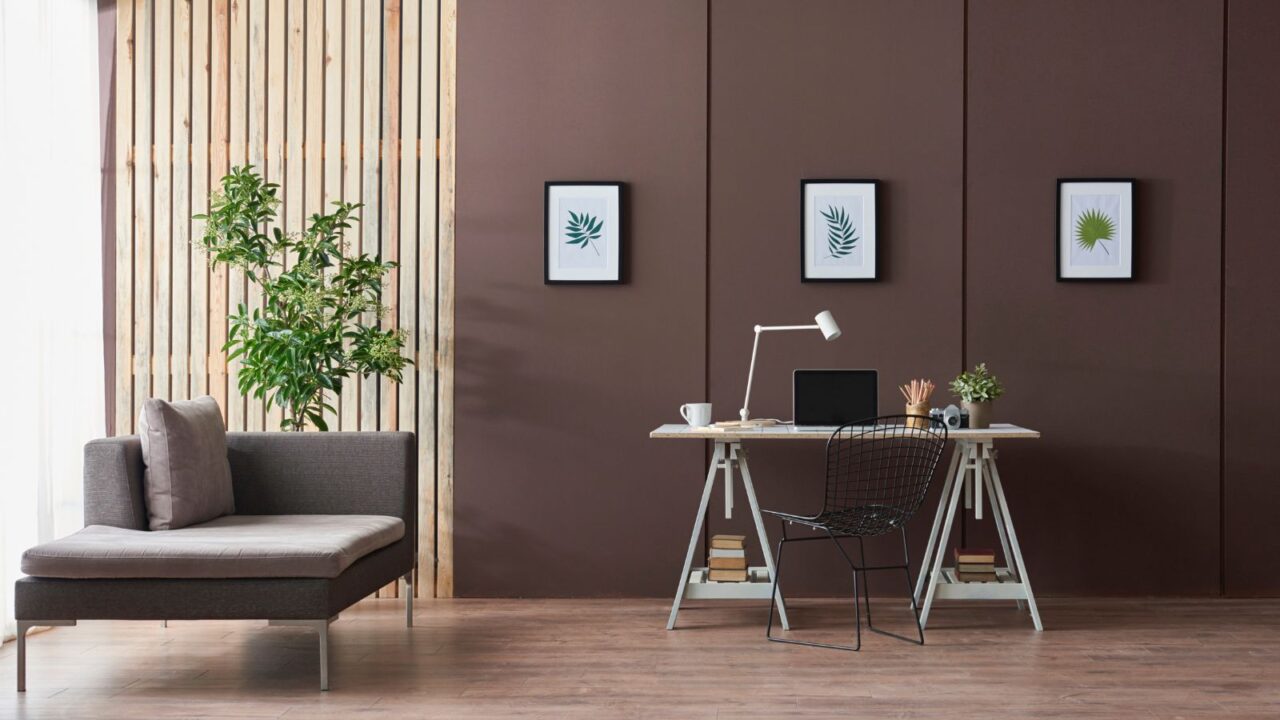 A minimalist home office with a table, chair, computer, lamp, frames, plant, and a sofa.