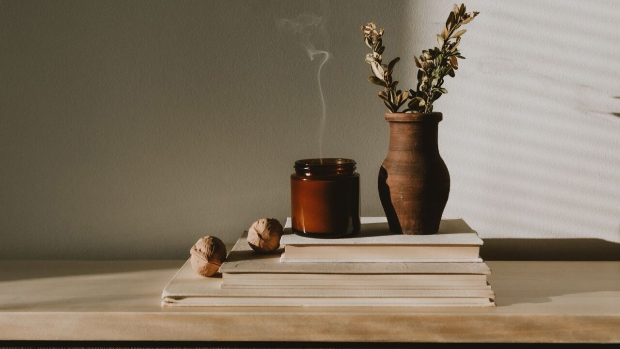 A wooden shelf with a layering of books, candle, nuts, and flower vase.