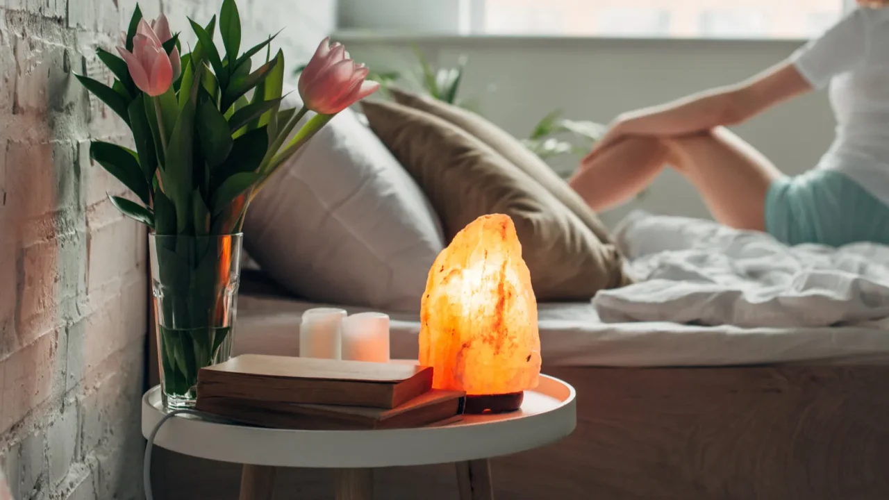 A round nightstand with books, Himalayan salt lamp, and lilies in a vase.