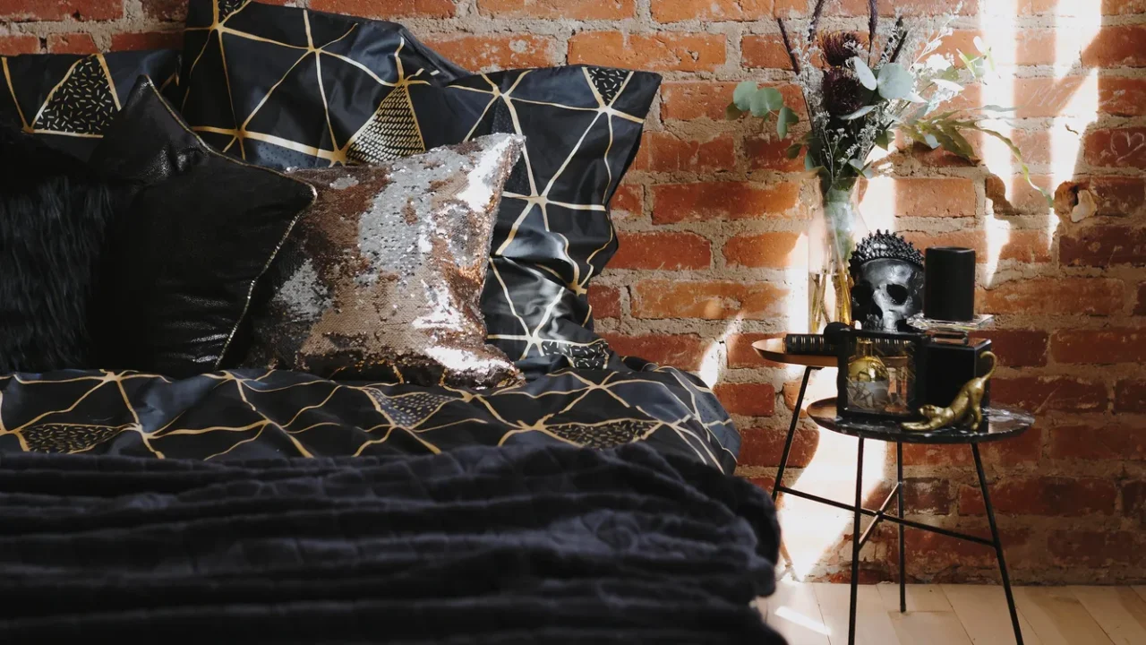 A bed in front of a brick wall with black and gold geometric patterned bedding, shimmery cushion, and bedside table with decor accents.