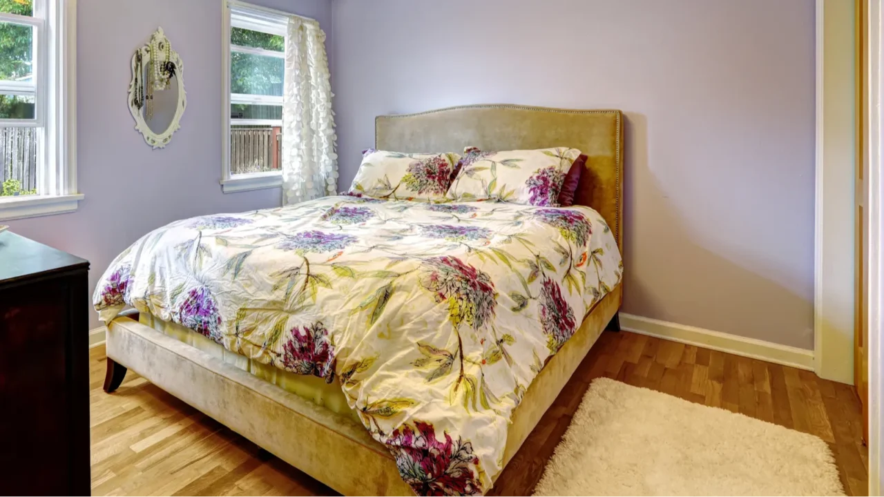 A bedroom interior with powdery lavender walls, wooden floor with rug, and a cozy bed.