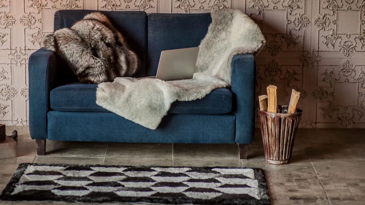 A living room interior with blue sofa with fur throw blanket, throw pillow, and laptop, firewood in rustic basket, and a shaggy black and white wool rug.