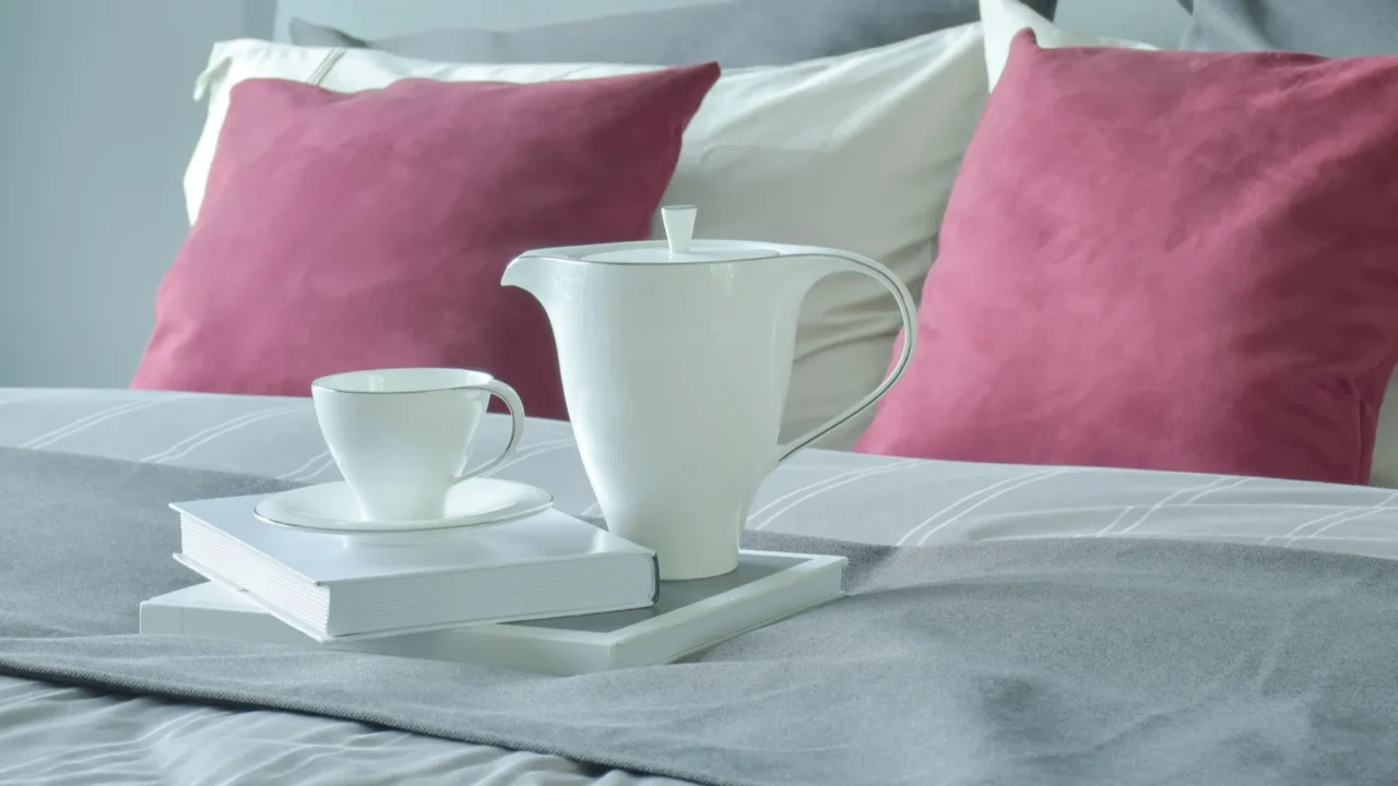 A bed with pink and white pillows, and a ceramic pot, cup with saucer, and books on a bed runner.