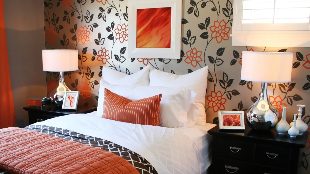 A cozy bedroom interior with white bedding, burnt orange cushion and comforter, side tables with lamp and vases, and framed artwork above the bed.