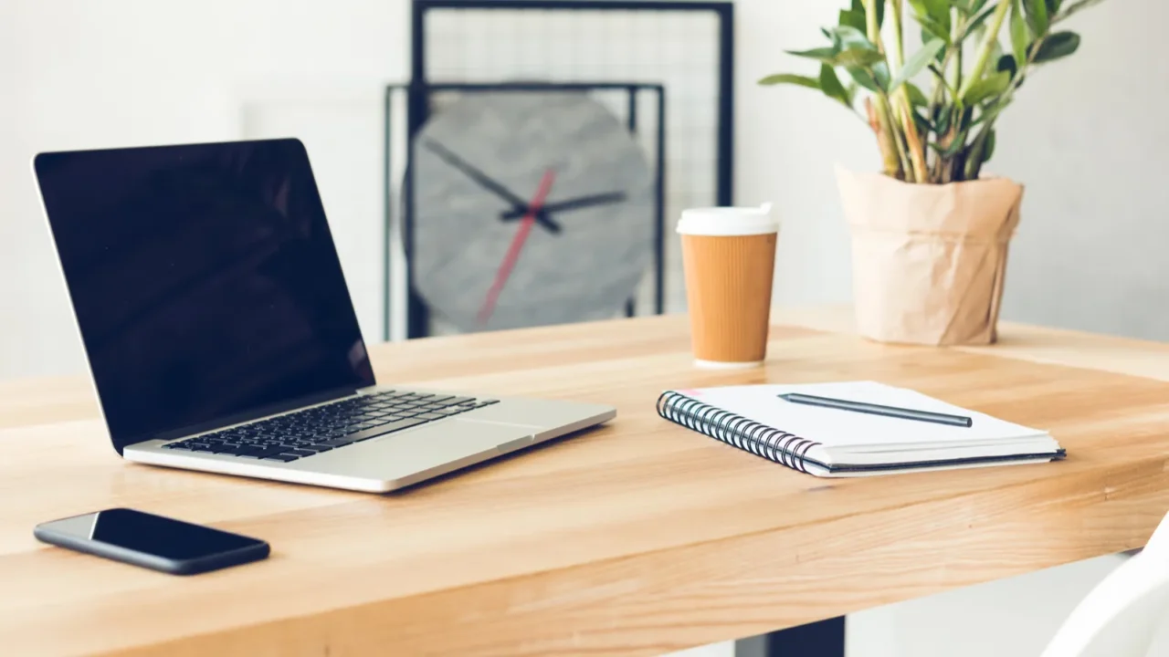 A wooden table with a laptop, phone, notepad, and a plant on it.