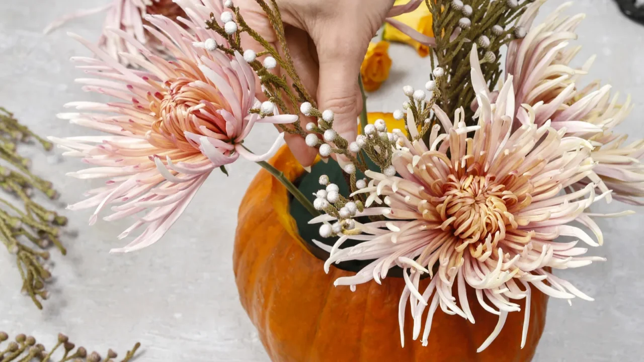 A person arranging flowers in a pumpkin placed on a white table.