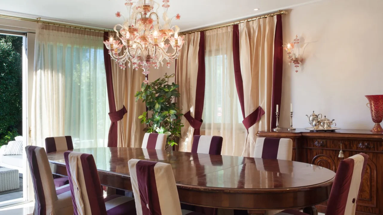 An elegant dining room featuring a large wooden table surrounded by chairs with cream and burgundy upholstery, draped with luxurious curtains, and adorned with a lavish chandelier.