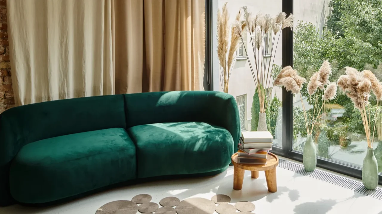 A green velvet sofa next to a window with pampas grass in vases and a stack of books on a wooden table.