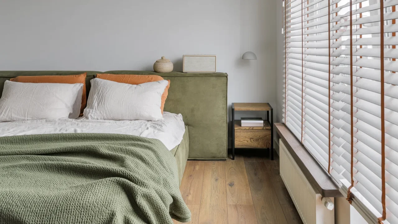 A modern japandi bedroom interior with moss green and white bedding, wooden nightstand, and roller blinds.