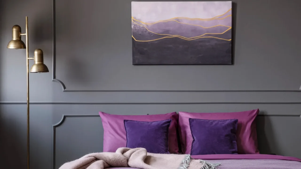 A stylish bedroom interior with violet bedding, velvet cushions, golden floor lamp, and abstract artwork on a gray wall.