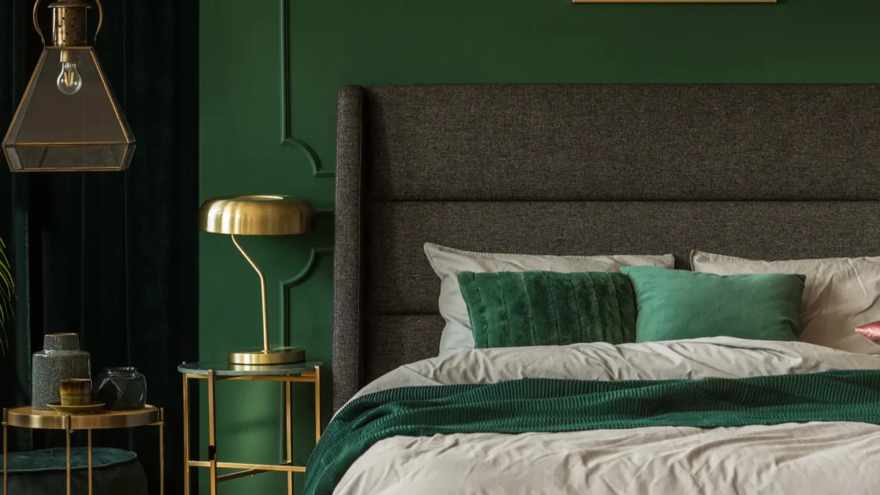 A modern emerald green bedoom interior with cozy bed, gold frame side table with lamp, and statement ceiling light.